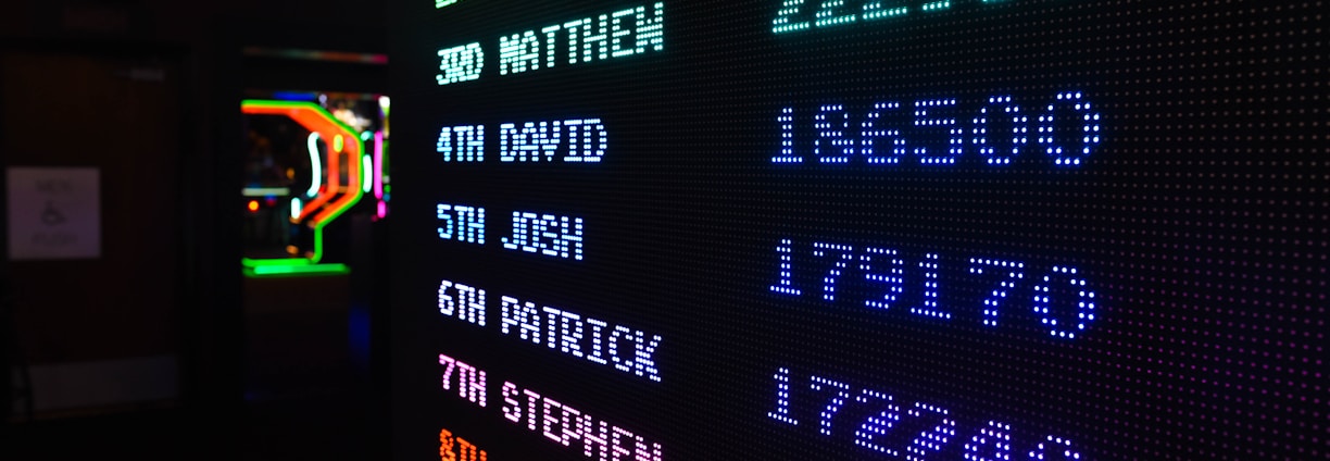 A digital leaderboard displaying names and scores in vibrant colors like green, blue, purple, and orange. The display is in a dark setting, possibly an arcade, and features a bright LED design.