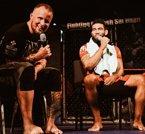 Two individuals are seated on a stage, each holding a microphone and smiling. They are in a sports-related setting with one wearing a casual black outfit and the other in sportswear, with a towel around his neck. The background includes elements of a fighting or martial arts cage.