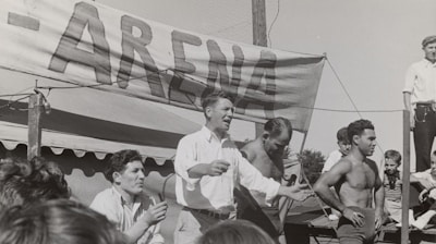 A group of men, some shirtless, are gathered around a platform or stage. A man in a white shirt appears to be speaking or gesturing to a crowd. A large sign with the word 'ARENA' is prominently displayed in the background. The scene seems to be set outdoors on a sunny day.