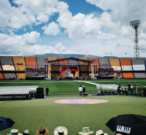 A sports stadium with a large stage set up on the field, hosting an outdoor event. Spectators wearing colorful hats sit in the foreground. The seating areas display vibrant colors, and a clear blue sky with scattered clouds is visible. Large screens display graphics related to the event.