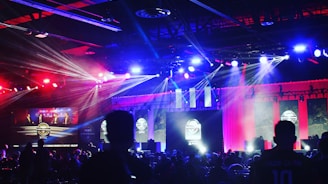 A dimly lit indoor arena filled with an audience. Bright stage lights in red and blue illuminate multiple large screens displaying graphics.