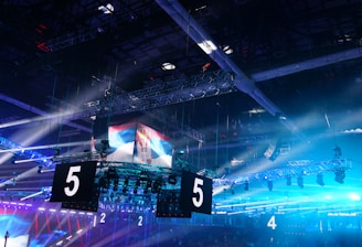 A large indoor arena is equipped with advanced lighting and audio systems. Suspended above the space are numerous digital displays showcasing numbers. The environment is filled with blue illumination and spotlights creating a vibrant atmosphere.