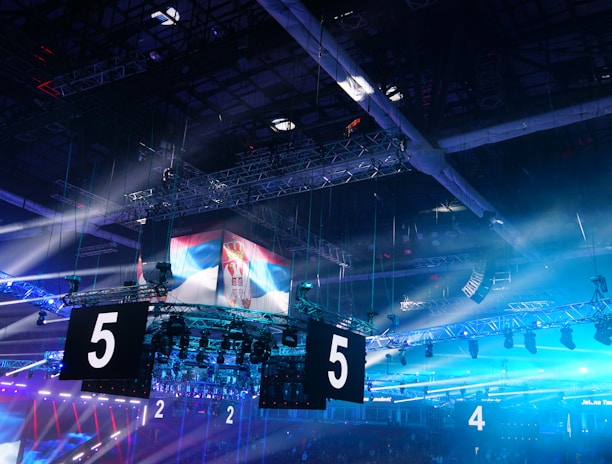 A large indoor arena is equipped with advanced lighting and audio systems. Suspended above the space are numerous digital displays showcasing numbers. The environment is filled with blue illumination and spotlights creating a vibrant atmosphere.