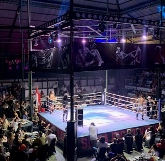 A crowded indoor arena with a boxing ring at the center, surrounded by an audience seated at tables. The scene is illuminated by overhead lights, casting a vibrant glow over the ring where two fighters are preparing for a match. Spectators are engaged, capturing the event with cameras and enjoying the lively atmosphere.