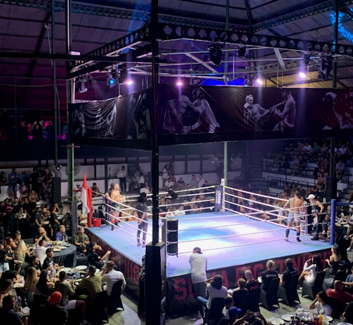 A crowded indoor arena with a boxing ring at the center, surrounded by an audience seated at tables. The scene is illuminated by overhead lights, casting a vibrant glow over the ring where two fighters are preparing for a match. Spectators are engaged, capturing the event with cameras and enjoying the lively atmosphere.