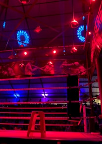 A brightly lit boxing or wrestling ring with vivid red and blue lights. Banners with images of fighters hang from the ceiling. The ropes of the ring are prominently visible, and a stool is placed inside the ring. The arena is filled with an audience, silhouetted against the colorful lighting.