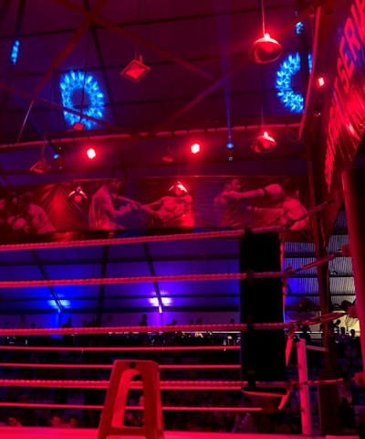 A brightly lit boxing or wrestling ring with vivid red and blue lights. Banners with images of fighters hang from the ceiling. The ropes of the ring are prominently visible, and a stool is placed inside the ring. The arena is filled with an audience, silhouetted against the colorful lighting.