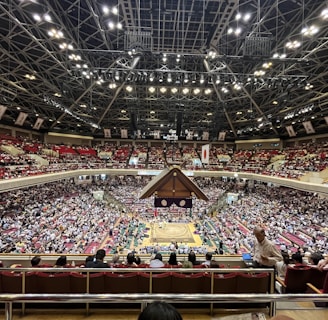 A large indoor arena packed with spectators seated in tiers around a central platform. The platform features traditional Japanese decor, indicating it may be used for sumo wrestling. The atmosphere is bustling, with people filling the seats and many spectators visible in the foreground.