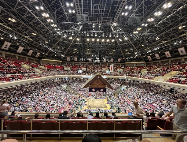 A large indoor arena packed with spectators seated in tiers around a central platform. The platform features traditional Japanese decor, indicating it may be used for sumo wrestling. The atmosphere is bustling, with people filling the seats and many spectators visible in the foreground.