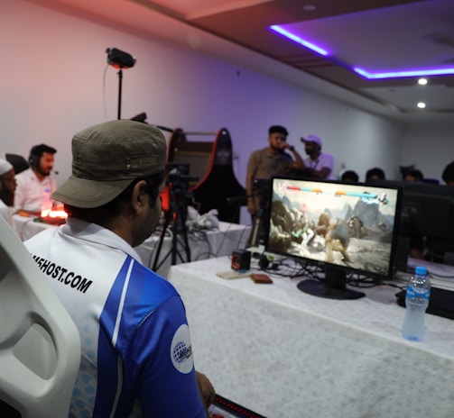 A group of people is gathered in a room watching and participating in a video game competition. Players and spectators are focused on the monitors displaying a fighting game. The setting includes tables with equipment and chairs arranged around the venue, with a noticeable intensity in the participants’ engagement.